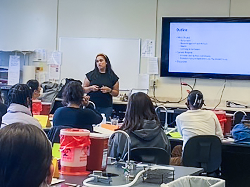 Cathy Samayoa, PhD, is standing and delivering a presentation on embodiment of chronic stress to a new cohort of SF BUILD scholars all seated in a lab, on July 5, 2022. 