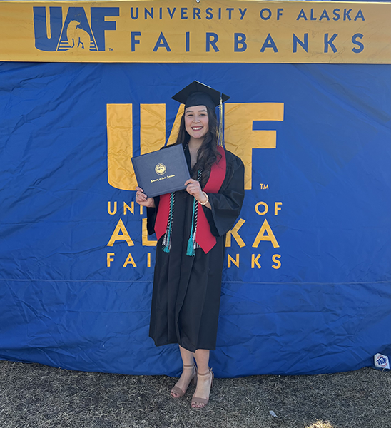 Samantha Wade is wearing her graduation cap and gown, standing in front of a UAF backdrop and posing with her degree. She earned a B.S. in Biological Sciences in May 2022.